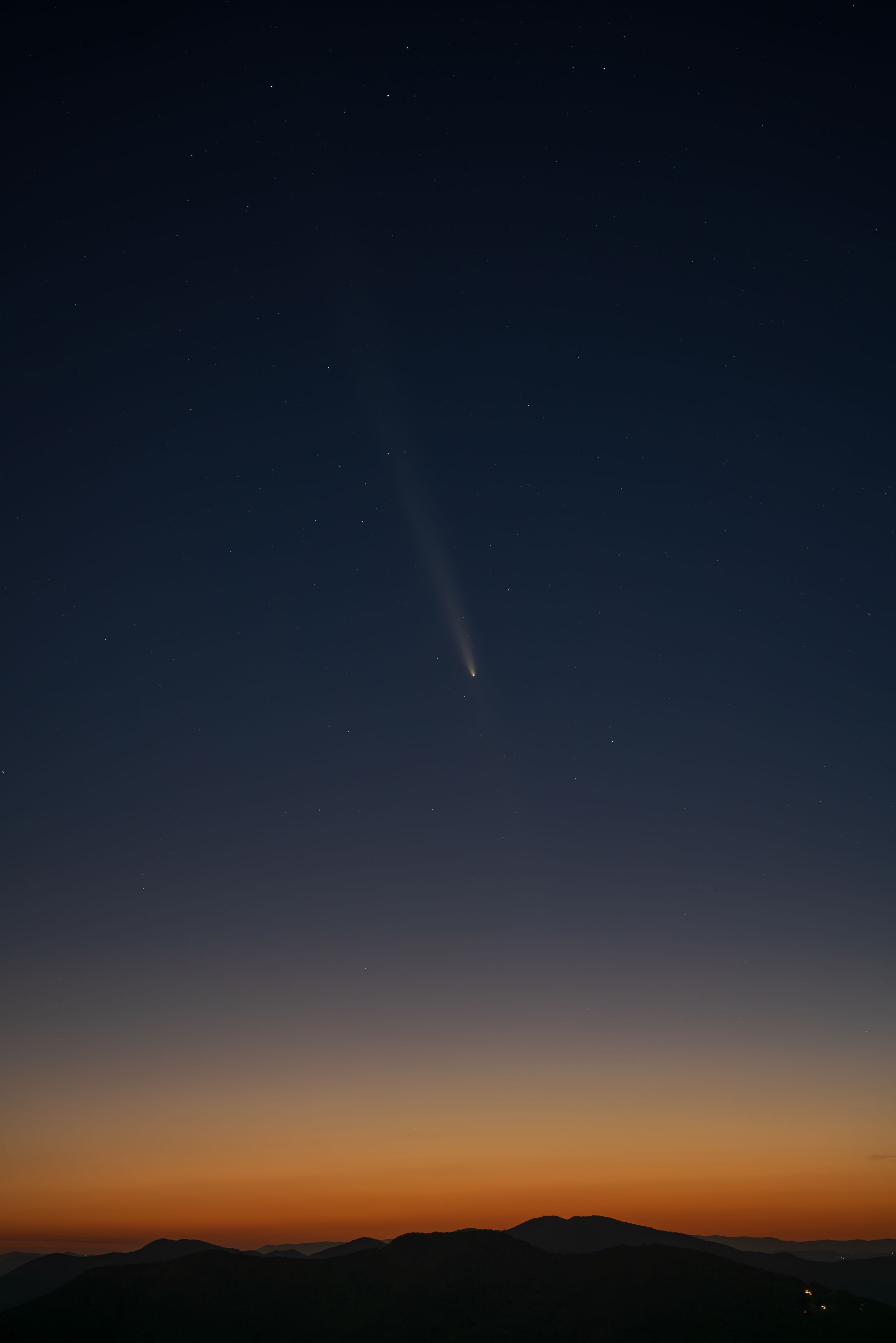 Comet streaking across a deep blue twilight sky above a dark mountain silhouette at dusk.