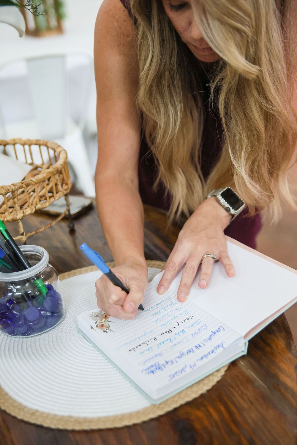Woman with blonde hair writing in a notebook with a blue pen at a wooden table with pens nearby.