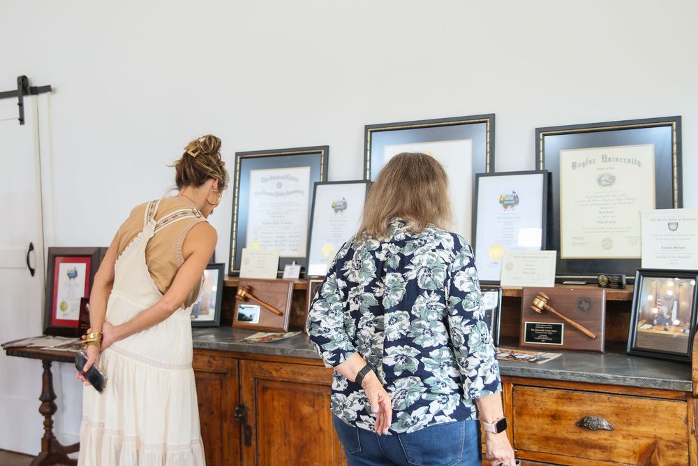 Two women viewing framed certificates and awards displayed on a wooden dresser at an indoor event.
