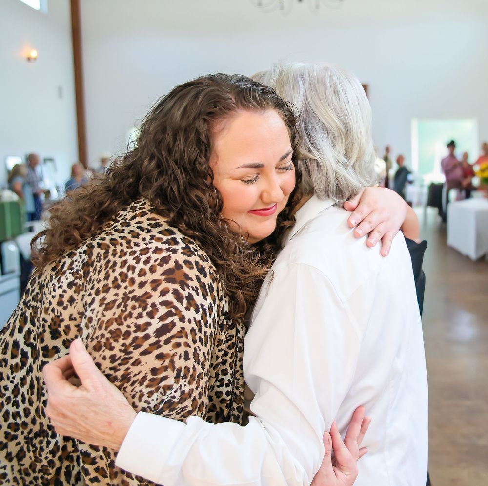 Two women sharing a warm embrace at an indoor event, one wearing a leopard print top, the other in white.