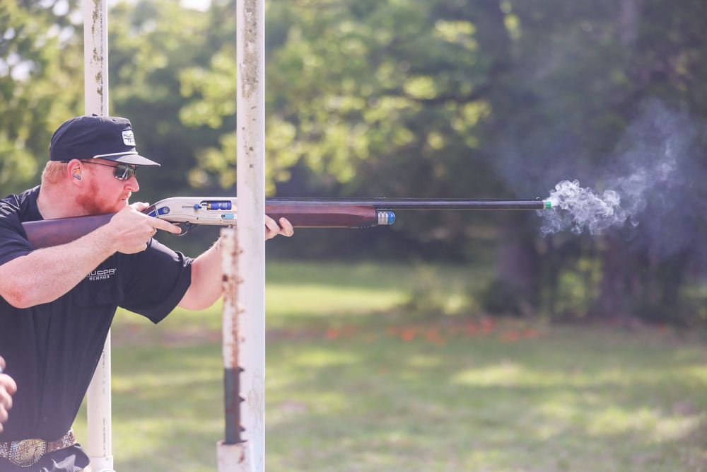 Man firing a long rifle outdoors at a shooting range with smoke visible from the muzzle.