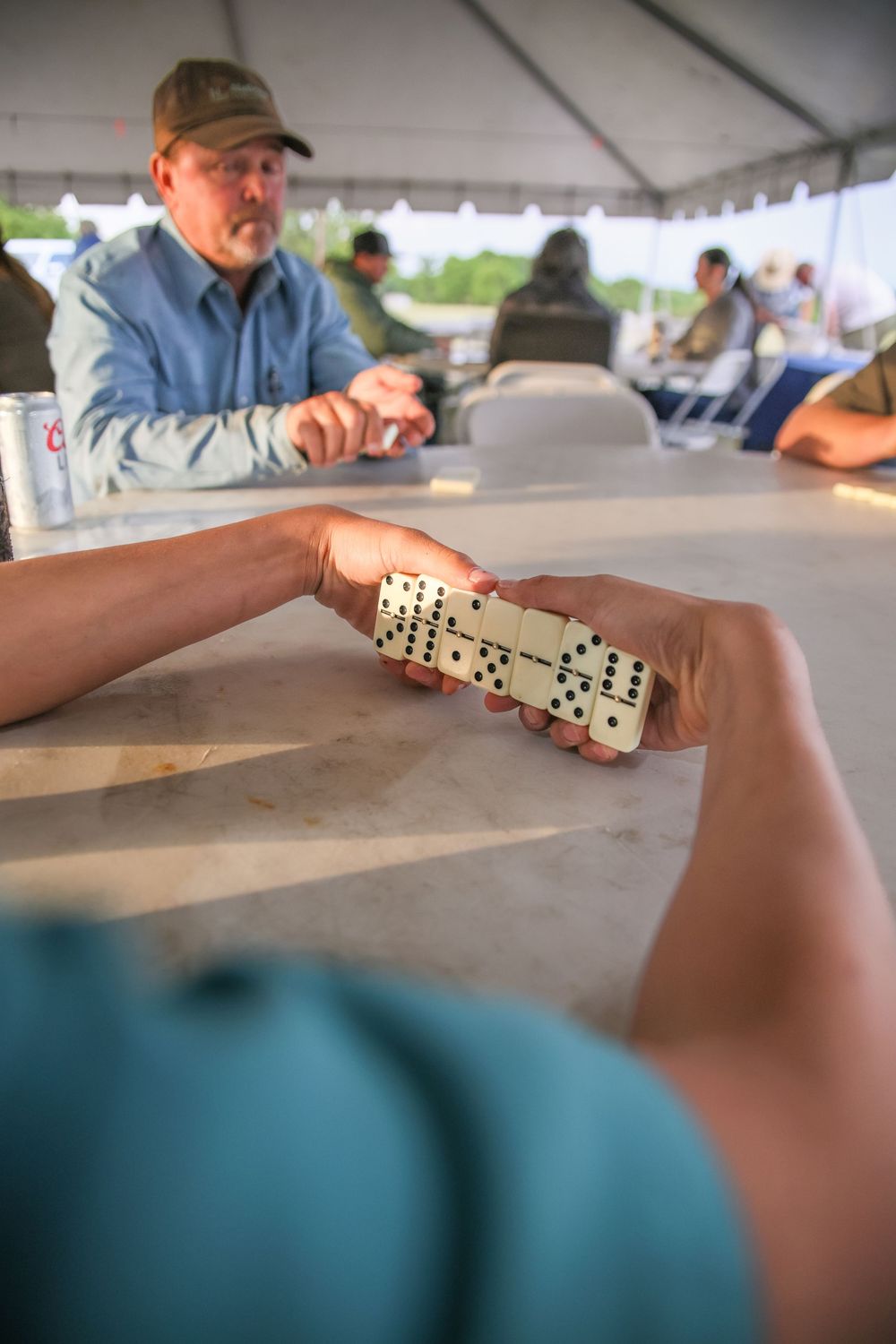 People playing dominoes under a tent, with a man in a cap visible across the table in the background.