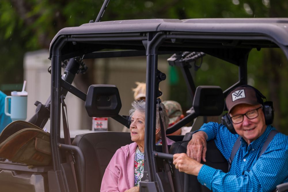 Elderly couple smiling in an off-road utility vehicle outdoors, man in blue shirt and headphones holding the steering wheel.