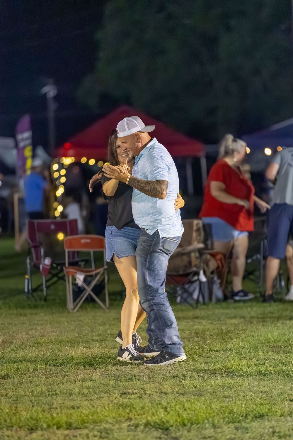 Couple dancing together at an outdoor evening festival on grass, with string lights and vendor tents in background.