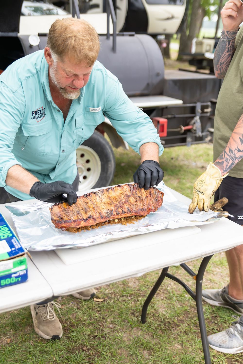 Man in blue shirt with black gloves slicing BBQ ribs on foil at outdoor barbecue competition near smoker.