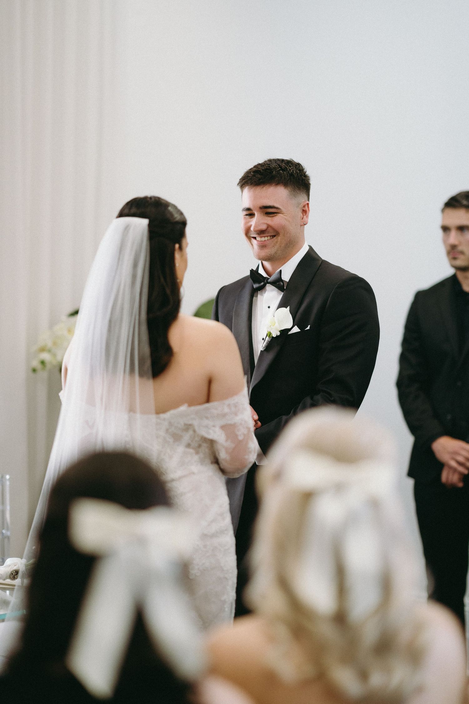 Groom smiling joyfully at bride during indoor wedding ceremony, wearing black tuxedo with white boutonniere.