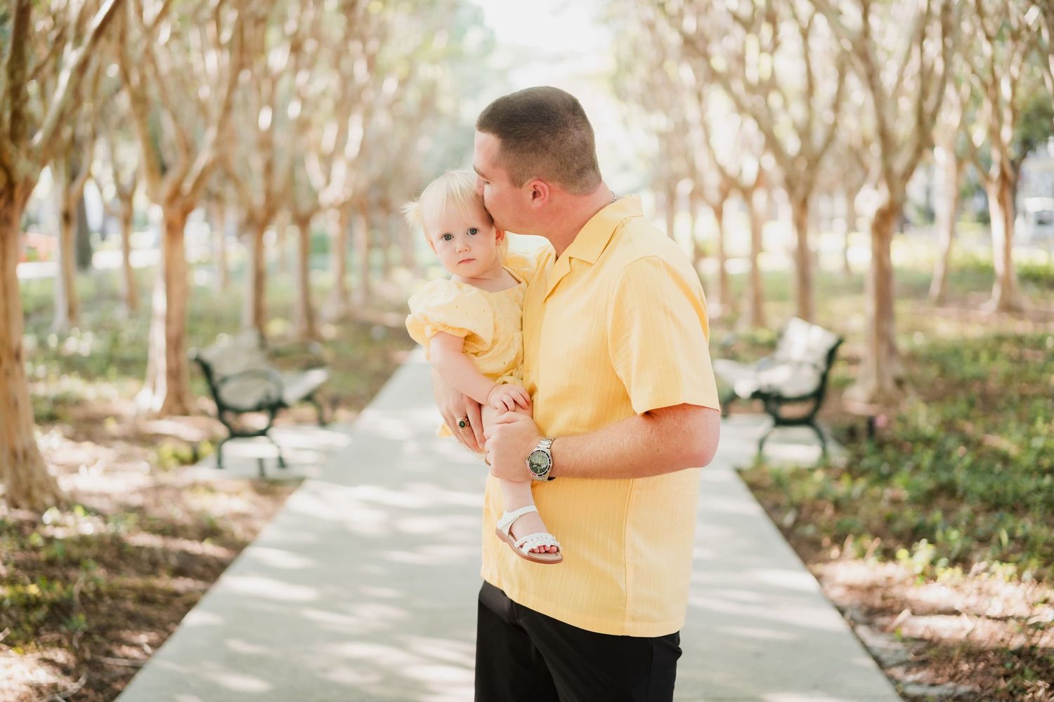 A father in a yellow shirt kisses his toddler daughter in a sunny park pathway lined with trees and benches.
