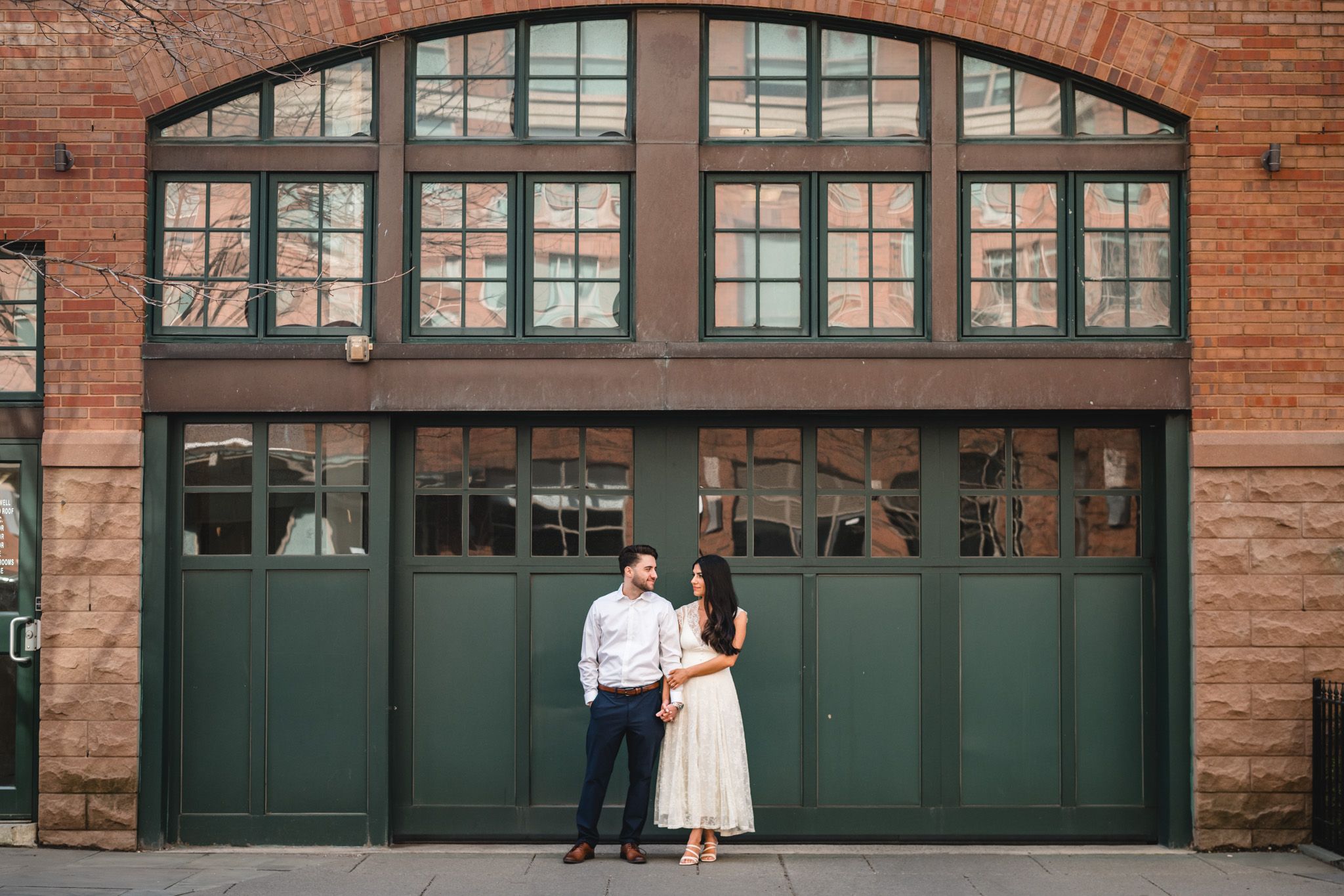Couple poses in front of green garage doors on a brick building for an engagement photo session.