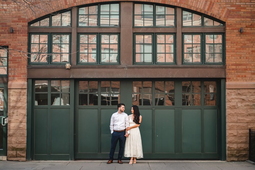 Couple poses in front of green garage doors on a brick building for an engagement photo session.