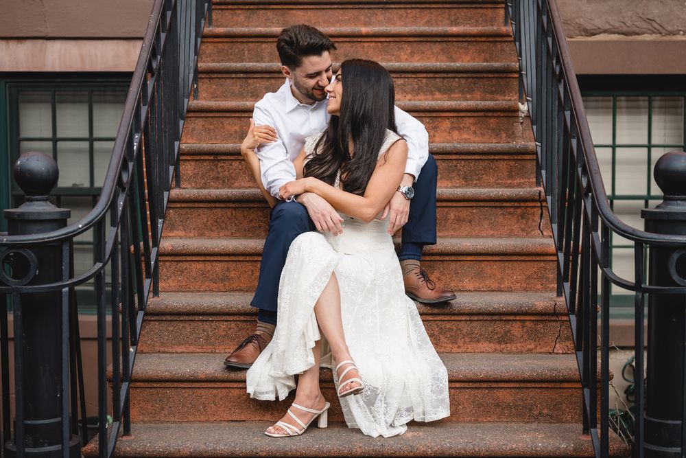 Couple sitting on brownstone steps, woman in white dress leaning into man in white shirt and navy pants.