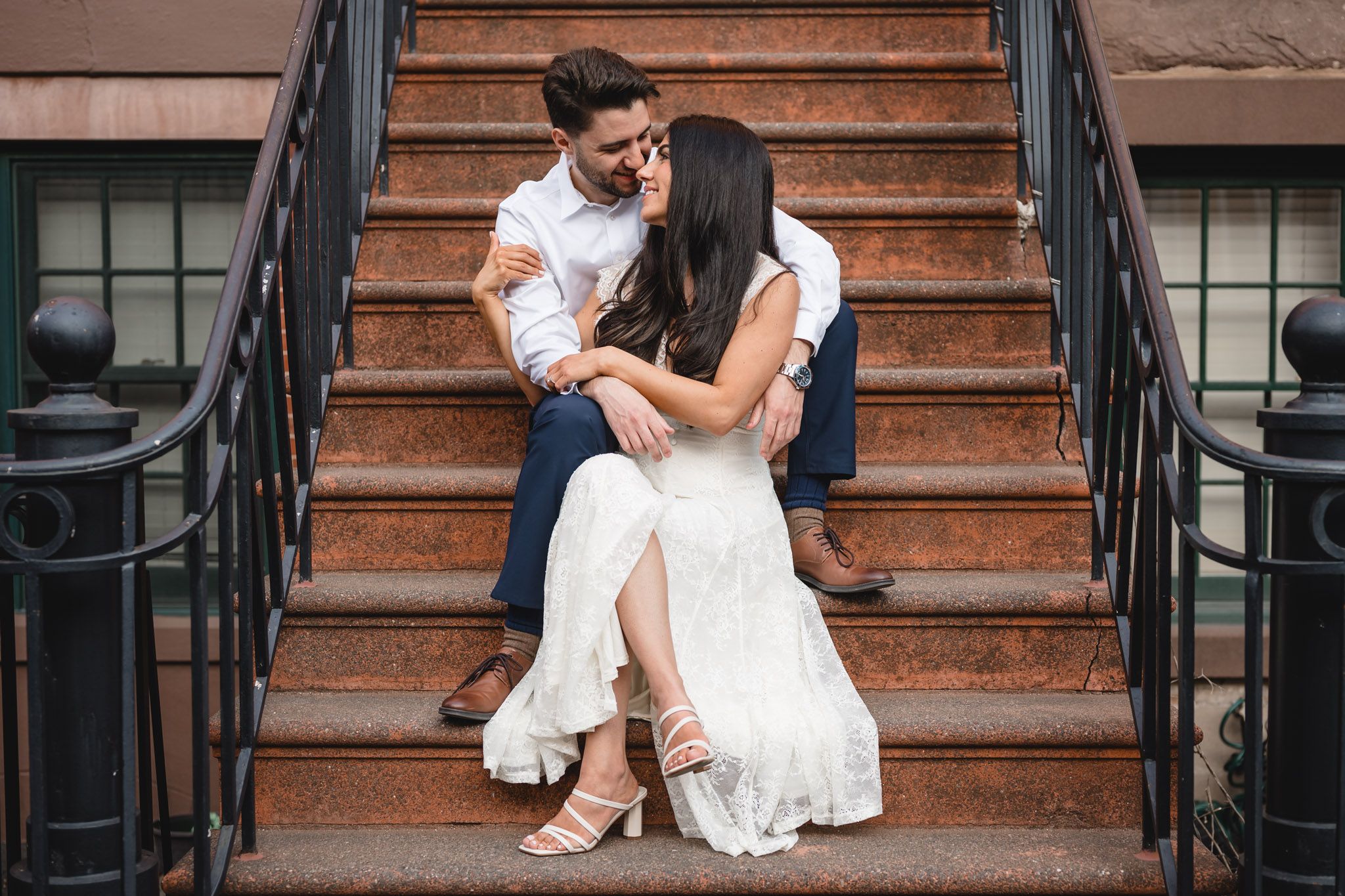 Couple sitting on brownstone steps, woman in white dress leaning into man in white shirt and navy pants.