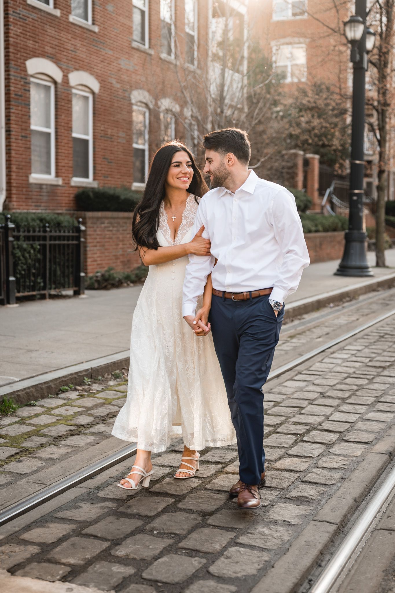 Couple walking hand-in-hand on cobblestone street, woman in white dress, man in white shirt and navy pants, brick buildings.