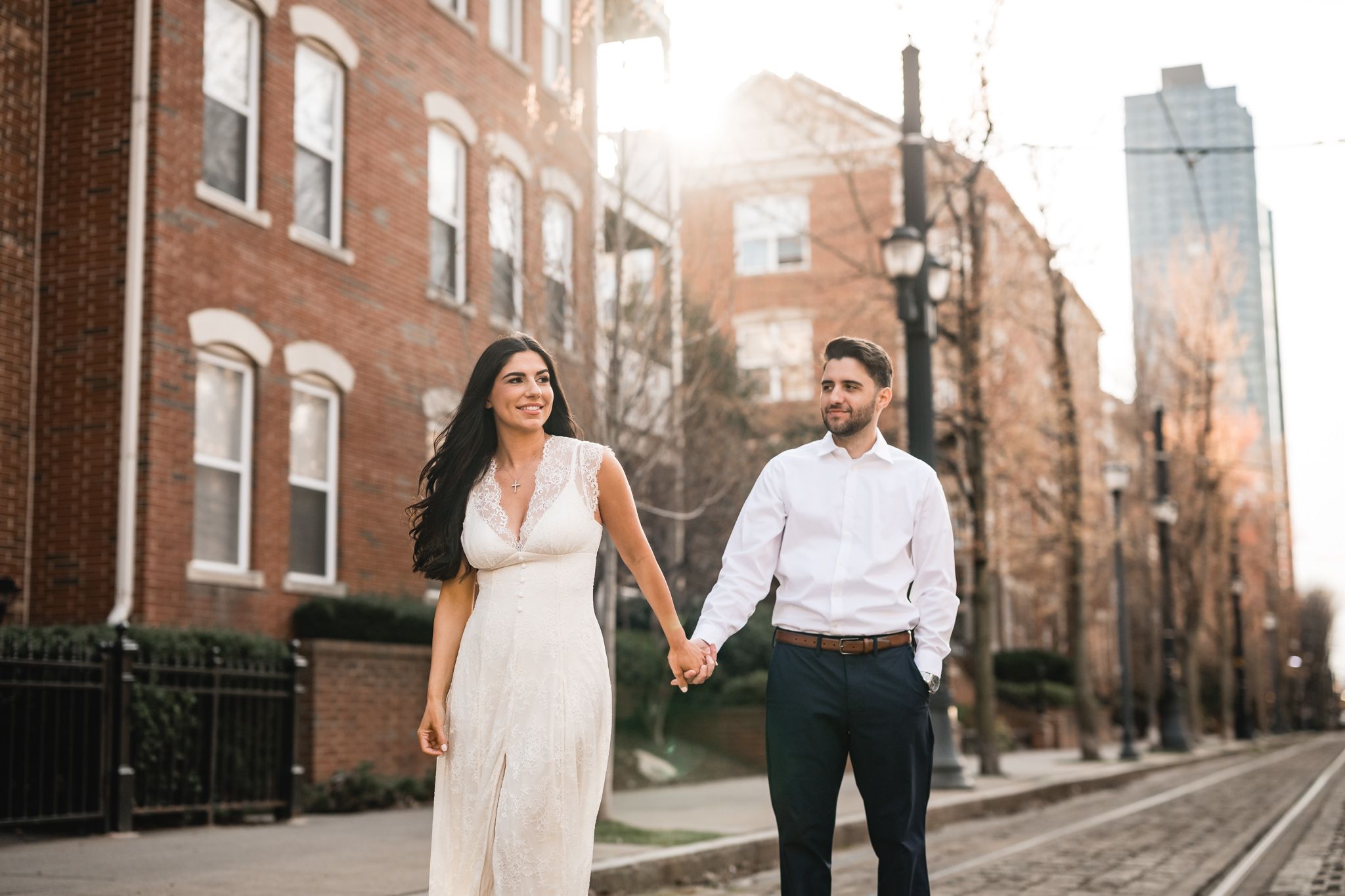 Couple holding hands strolling on city sidewalk with sunlight glowing behind brick buildings.