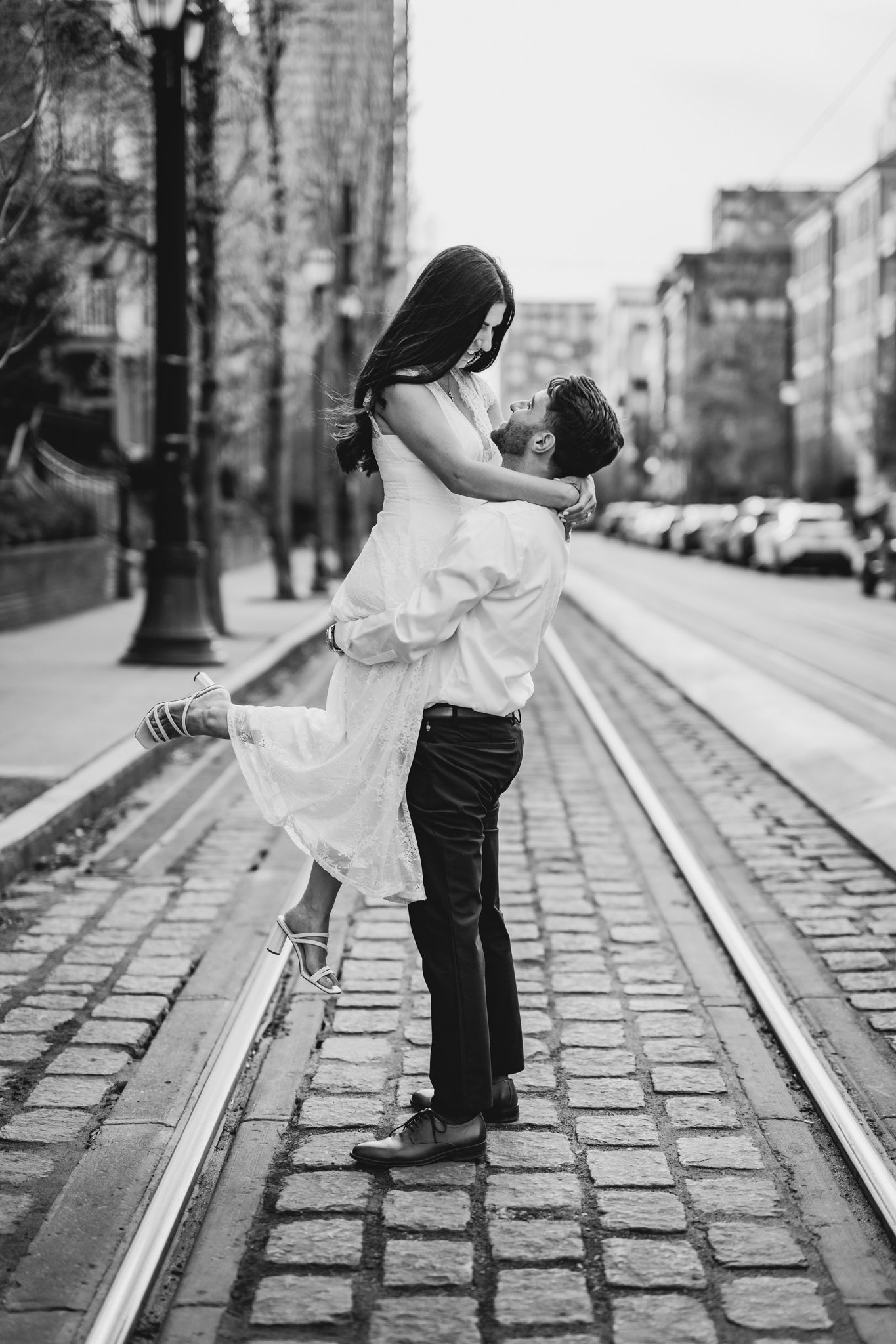 Black and white photo of a man lifting a woman in a white dress on a cobblestone street in the city.