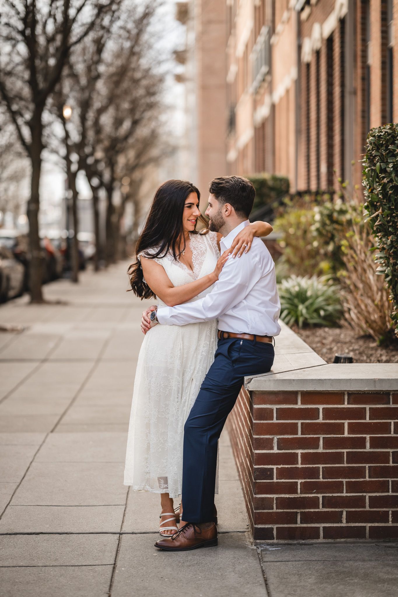 A couple in wedding attire embraces on a city sidewalk, the bride in white lace and groom in navy pants and white shirt.