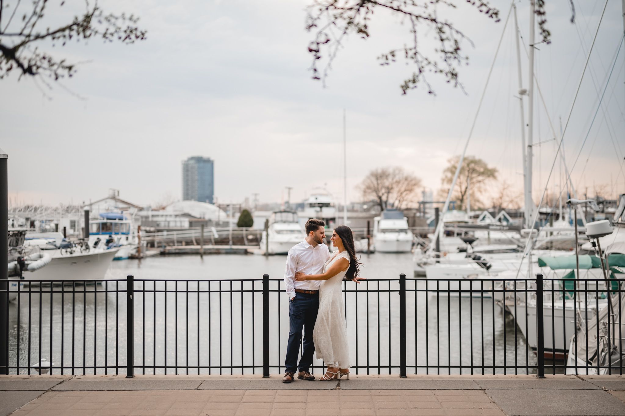 Couple embracing at a marina waterfront with sailboats and city skyline in the background during golden hour.