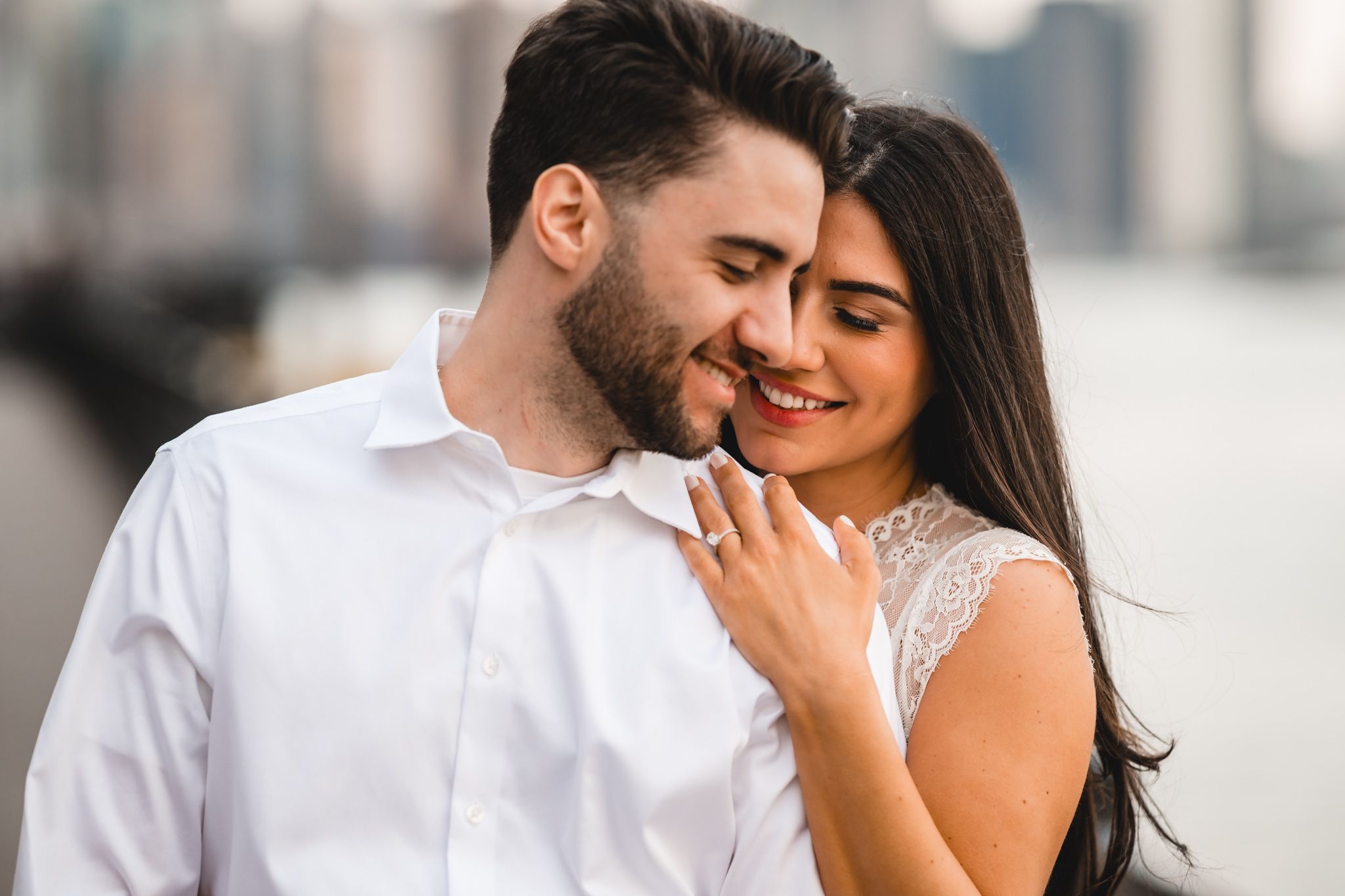 Happy couple embracing outdoors, woman showing engagement ring, city skyline in background.