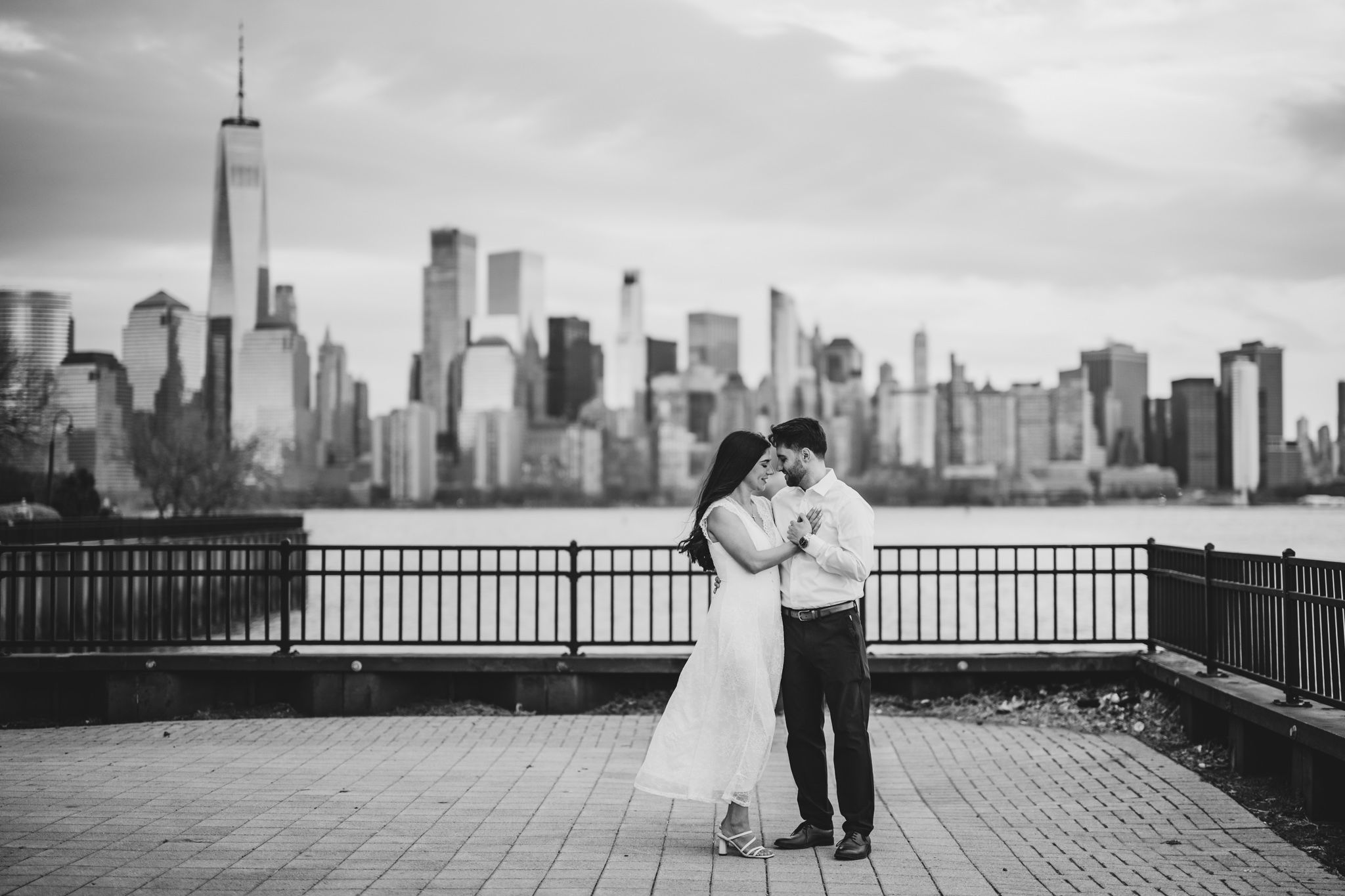 Couple embracing on waterfront promenade with NYC skyline and One World Trade Center in black and white.