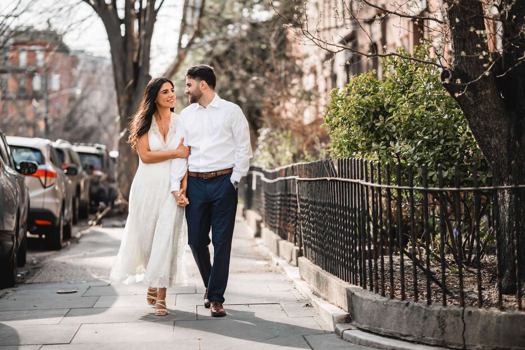 A couple walks hand-in-hand on a sunny urban sidewalk, smiling at each other in elegant white and navy attire.