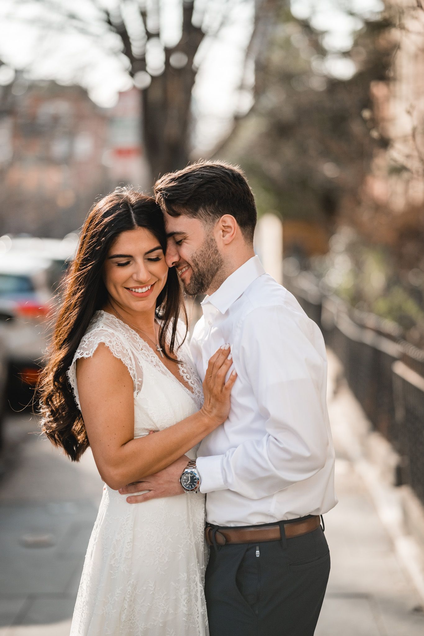 A smiling bride in a white lace dress embraces her groom in a white shirt on a sunny urban sidewalk.