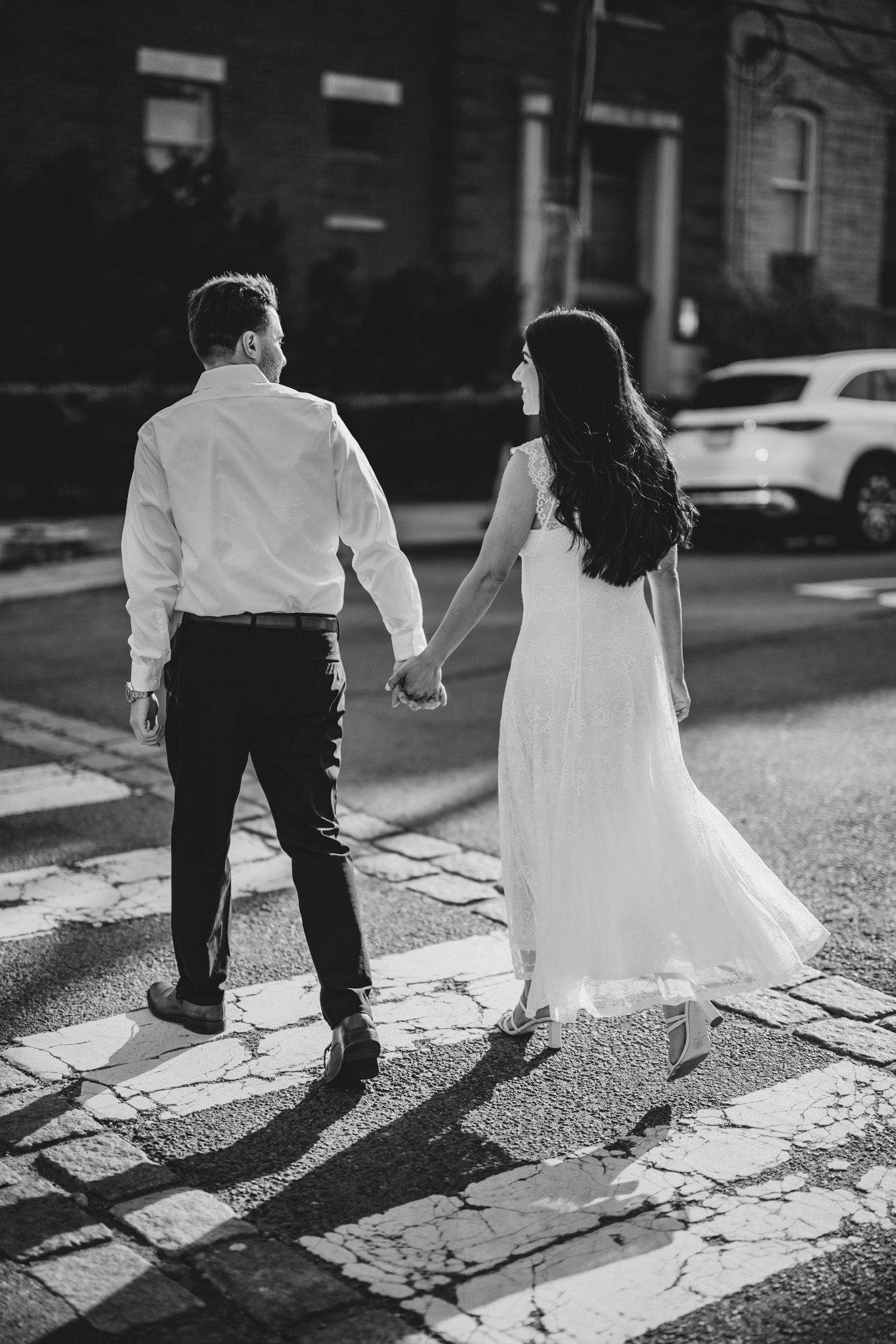 Black and white photo of a couple holding hands crossing a street, woman in white dress, man in white shirt and black pants.