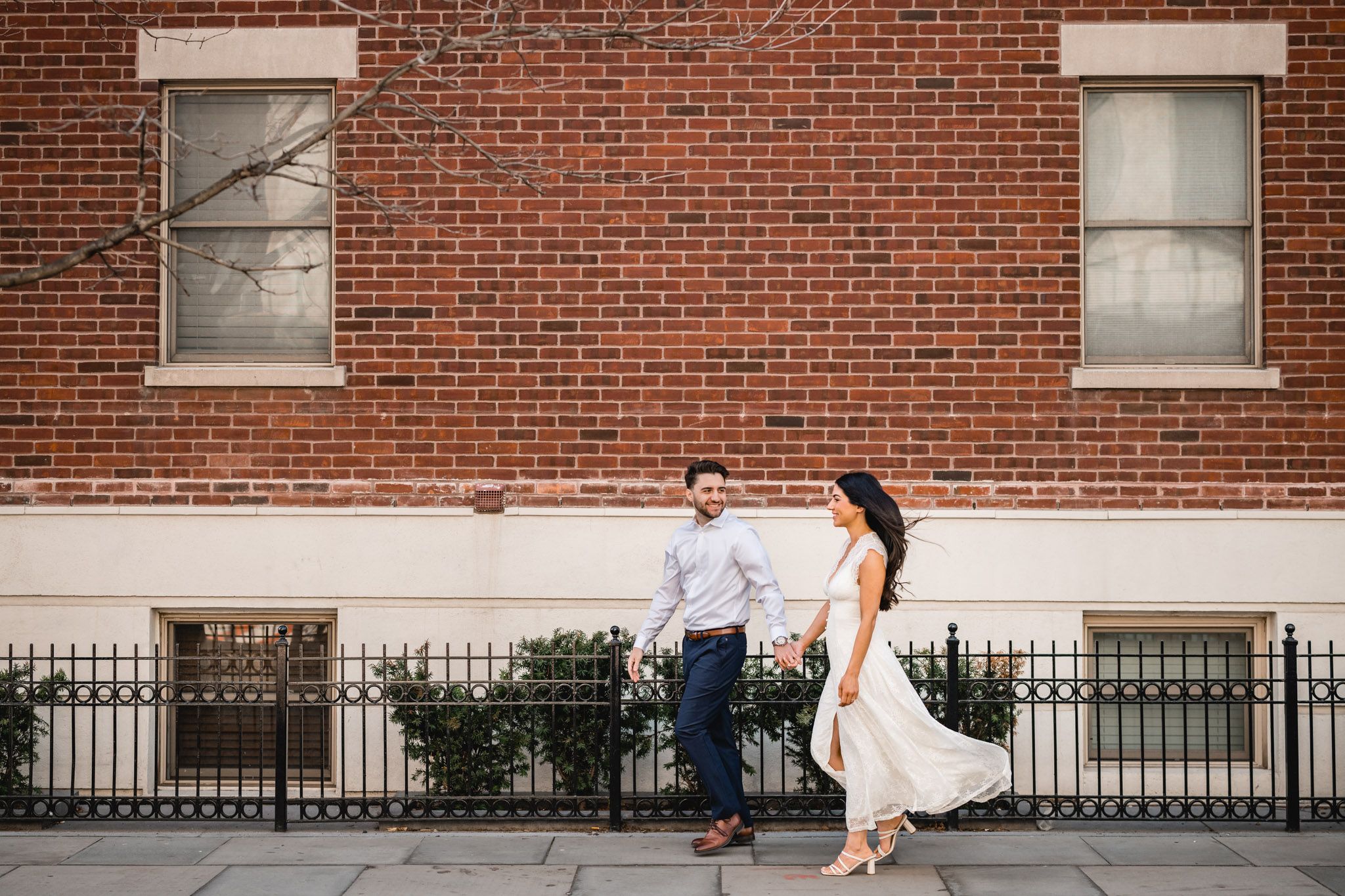 Couple holding hands walking along a brick building sidewalk, woman in white dress and man in blue pants and light shirt.