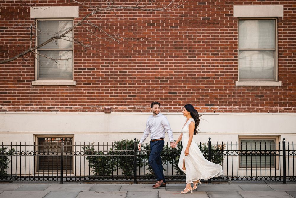Couple holding hands walking along a brick building sidewalk, woman in white dress and man in blue pants and light shirt.