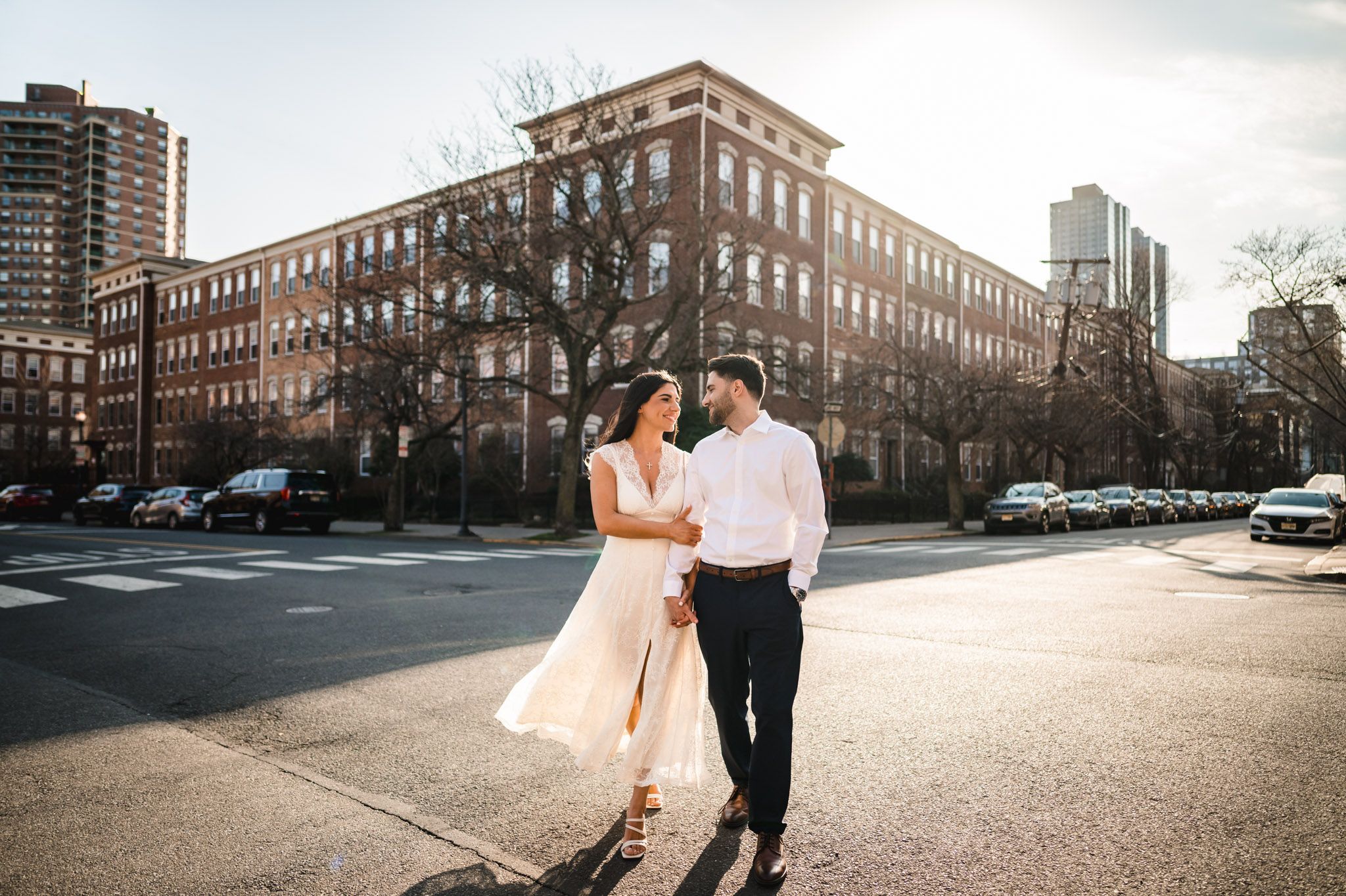 Couple walks hand-in-hand across urban street intersection with historic brick building backdrop on sunny day.