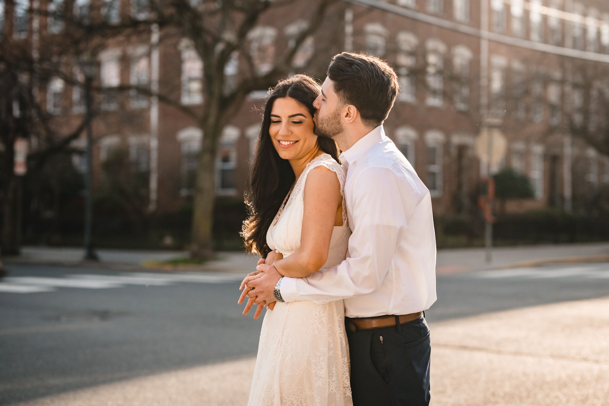 Couple embracing on a sunlit street, woman in white dress smiling as man kisses her cheek, warm golden hour light.