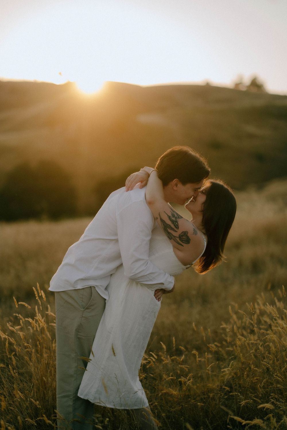 Couple sharing a romantic kiss in a golden field at sunset, with rolling hills in the background.