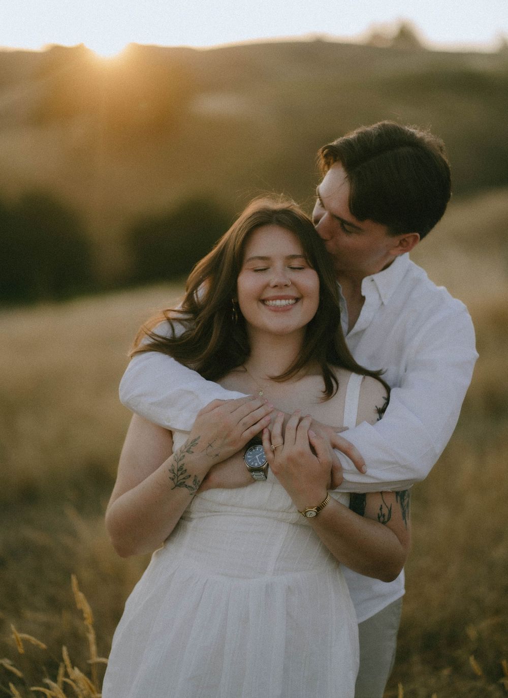 Couple embracing at golden hour in a field, woman in white dress smiling as man kisses her cheek.