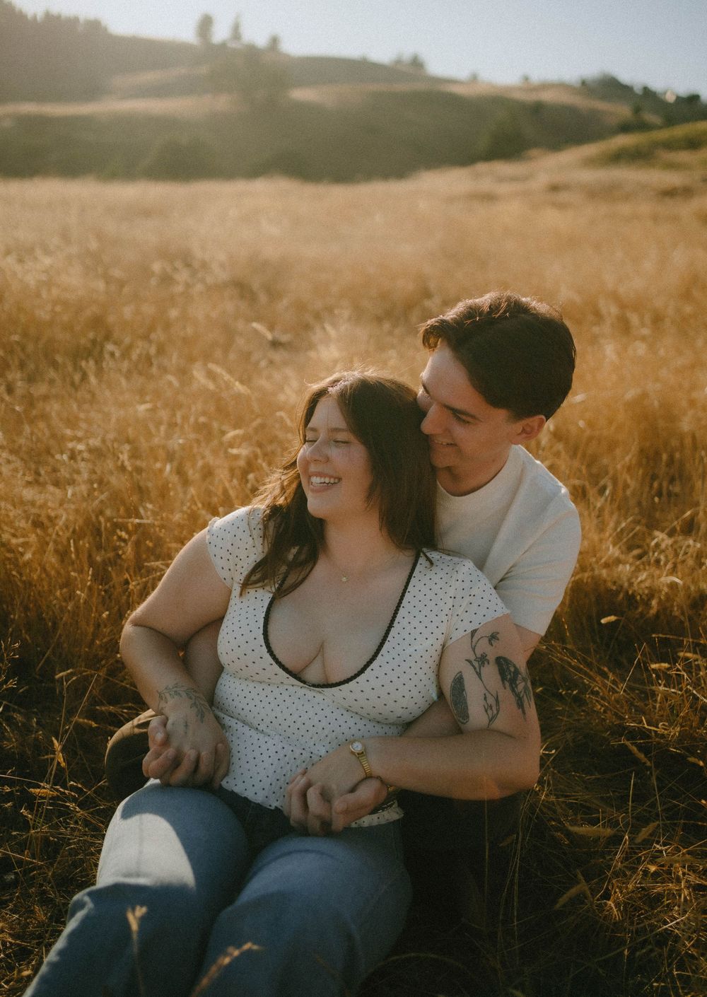 Couple sitting in golden field at sunset, woman laughing as man embraces her from behind.