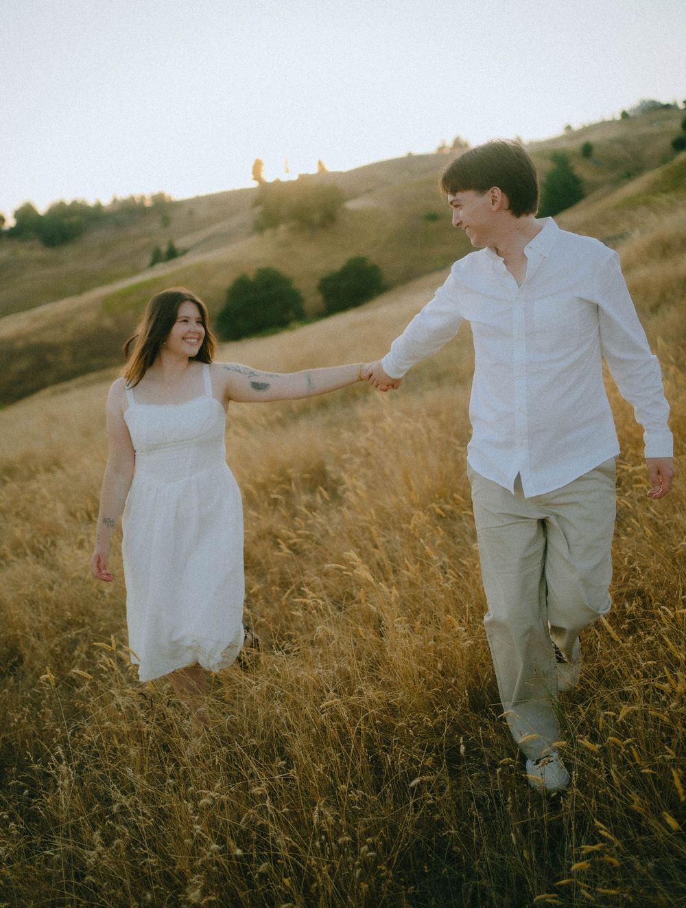 A couple holds hands walking through golden fields at sunset, woman in white dress, man in white shirt and khaki pants.