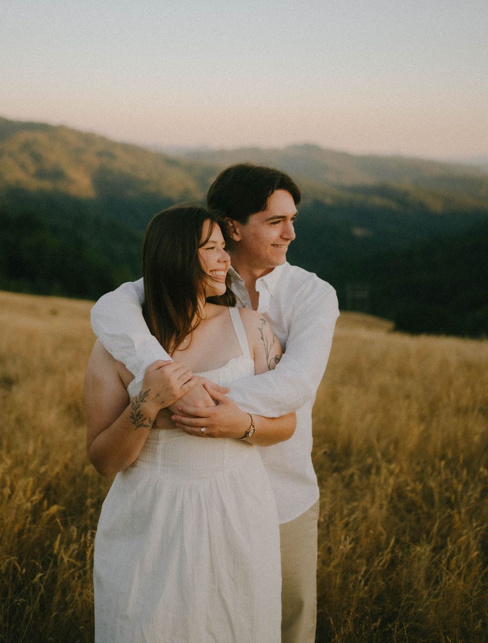 Couple embracing in golden field at sunset with rolling green hills in background, wearing white outfits.