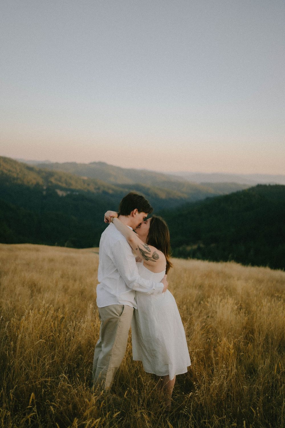 Couple kissing in golden meadow with mountain backdrop at dusk, wearing white outfits.