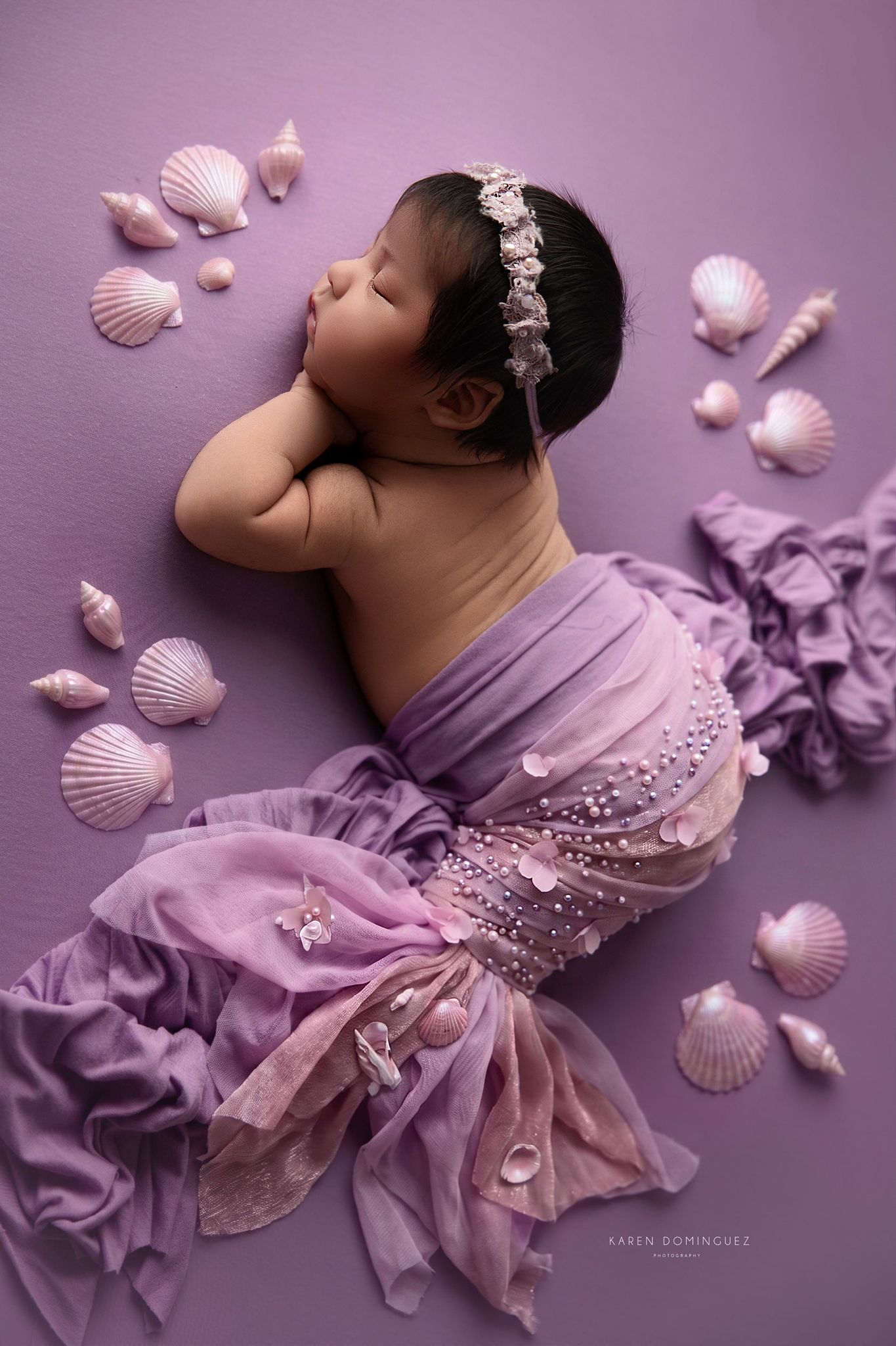 Newborn baby posed as a sleeping mermaid in purple fabric with seashells on a lavender background.