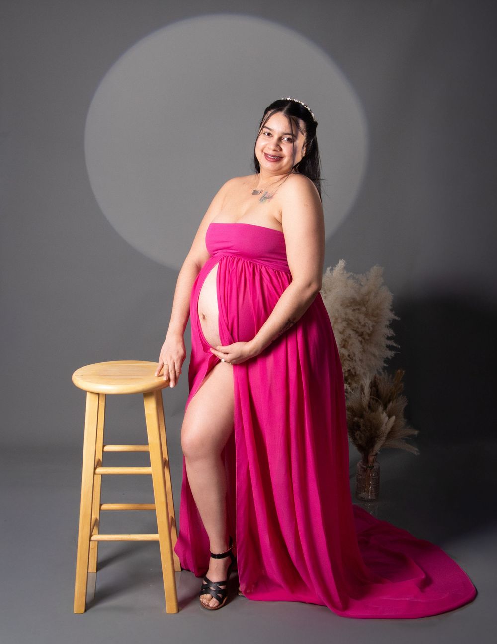 Pregnant woman in a flowing magenta gown poses gracefully beside a wooden stool in a professional studio setting.
