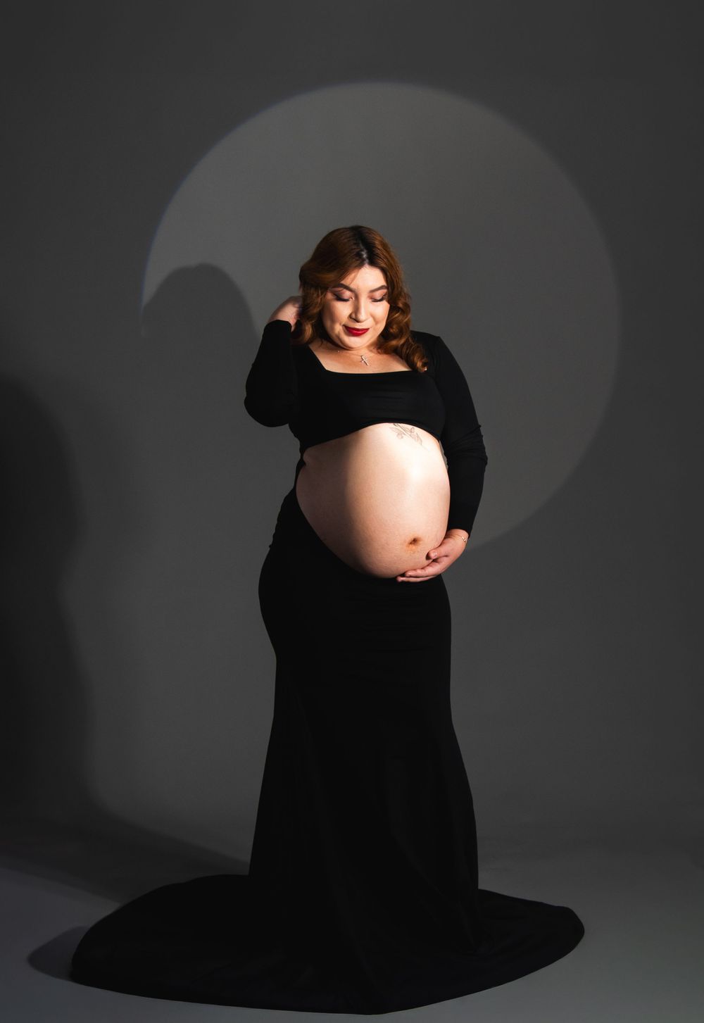 Pregnant woman in black maternity gown cradling bare baby bump during professional studio photo session.