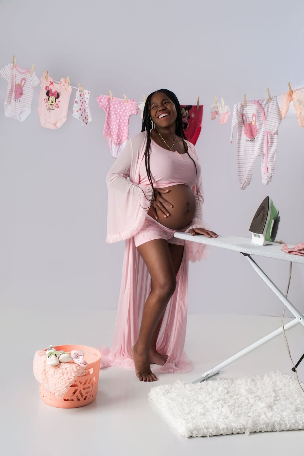 Smiling pregnant Black woman in pink robe touching baby bump near ironing board with baby clothes hanging in background.