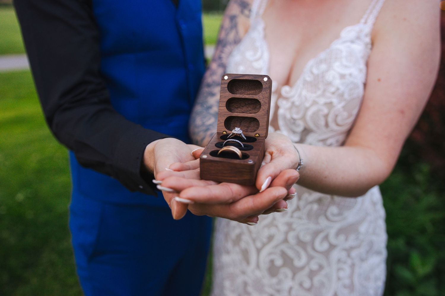 Wedding couple holding a rustic wooden ring box together in their hands against a green outdoor backdrop.