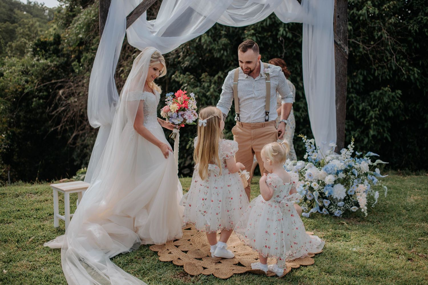 An outdoor wedding ceremony under a white draped arch with floral arrangements.