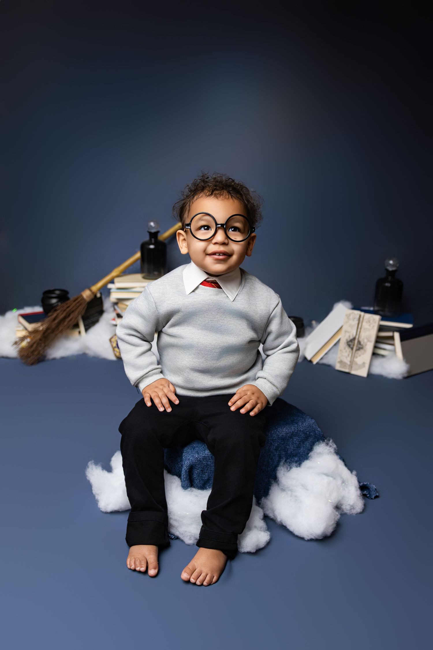 A toddler sitting on a blue backdrop decorated with clouds and items from Harry Potter. The toddler is wearing a grey sweater and glasses