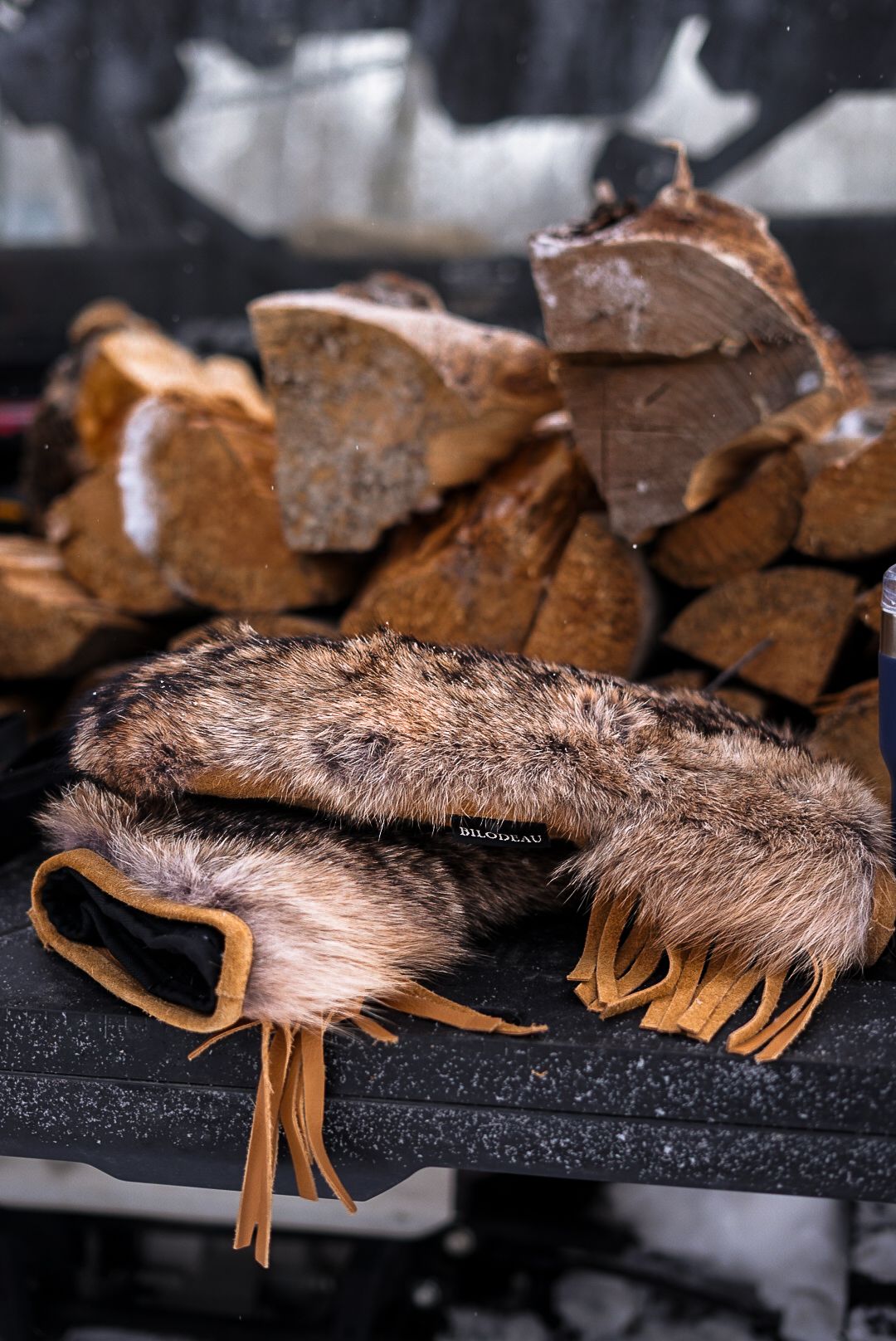 Furry winter accessories with leather fringe displayed against stacked firewood background.