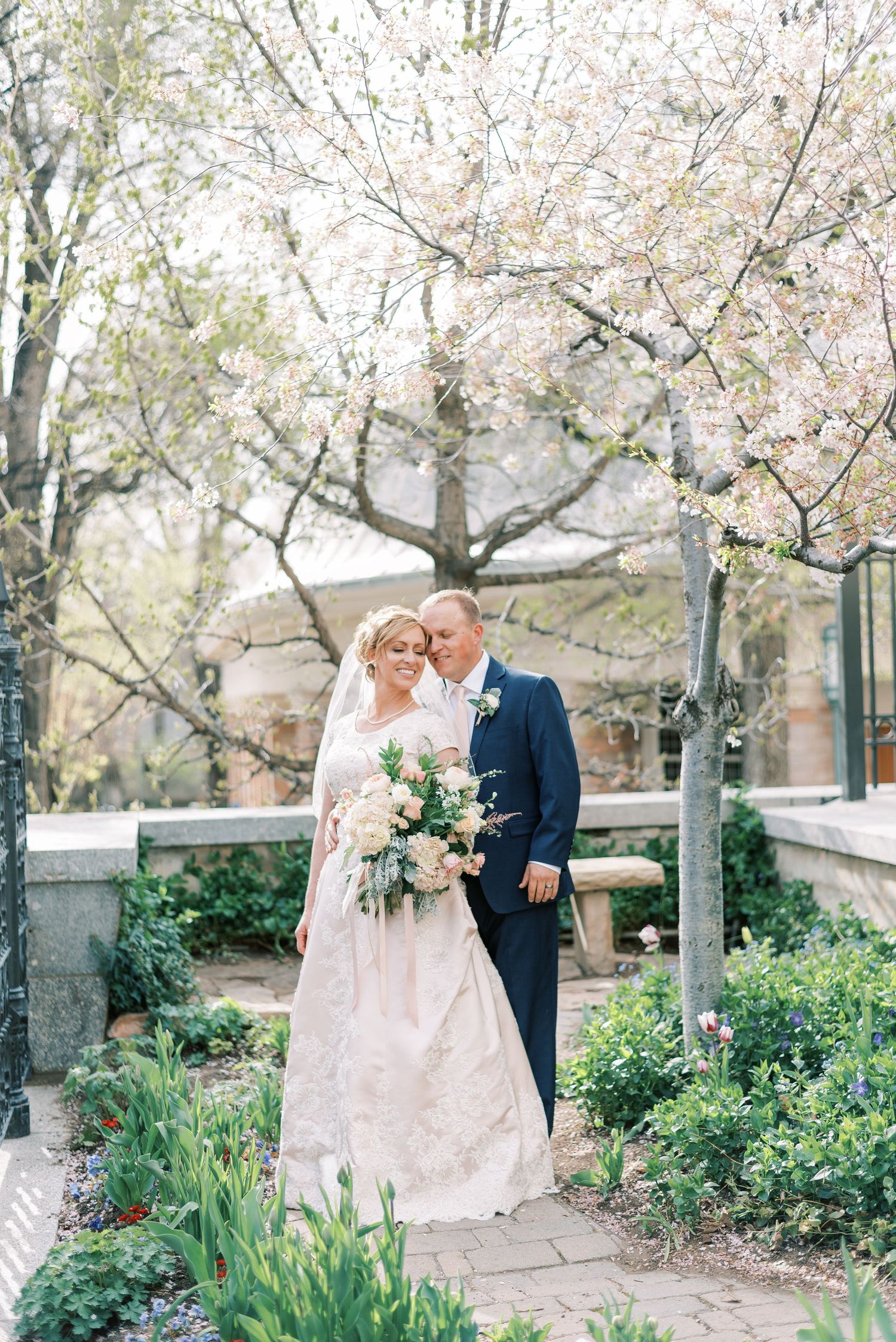 A springtime garden wedding scene with cherry blossoms blooming overhead as a couple embraces on a stone path.