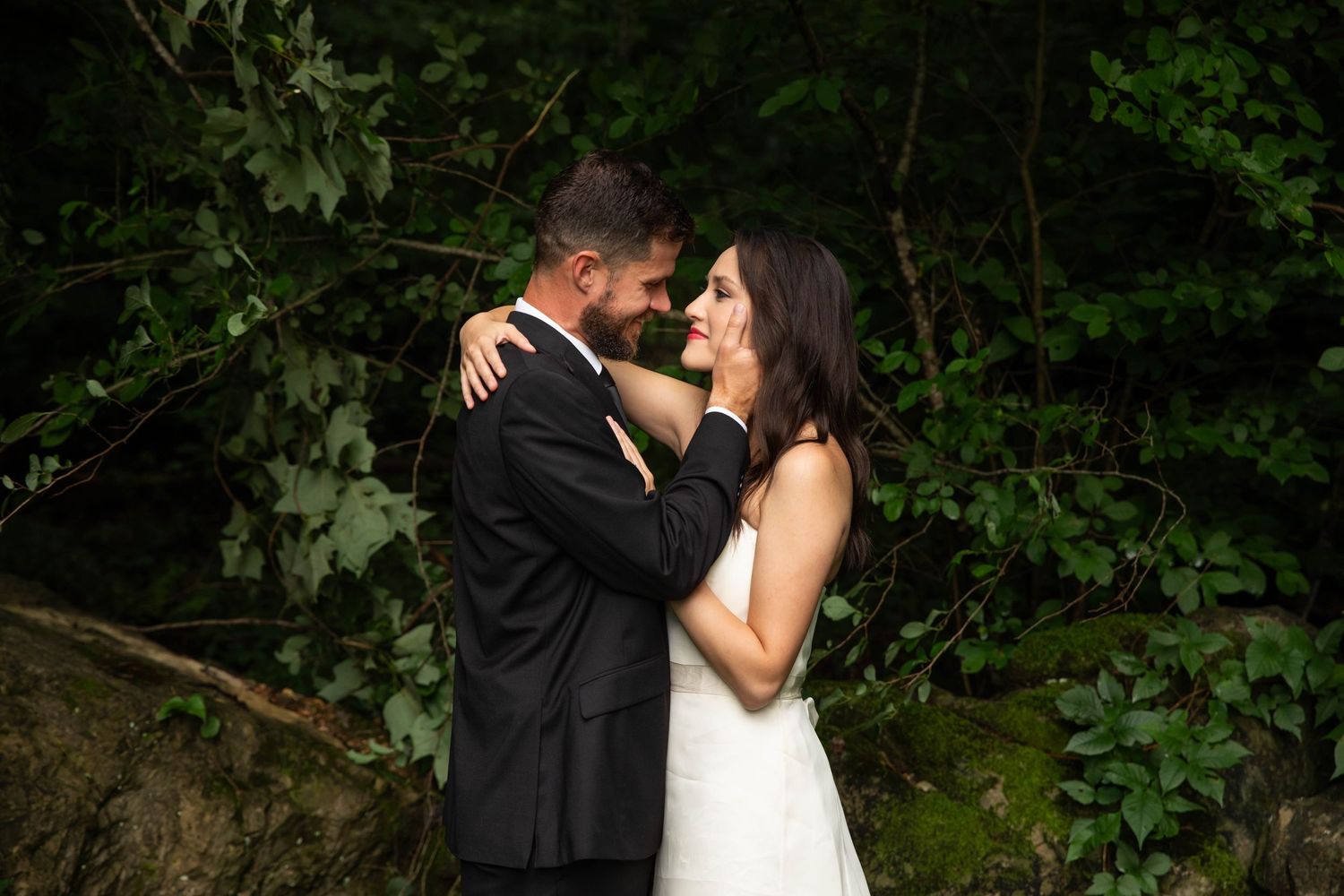 Photo of a bride and groom standing in the woods with his hand on her face.