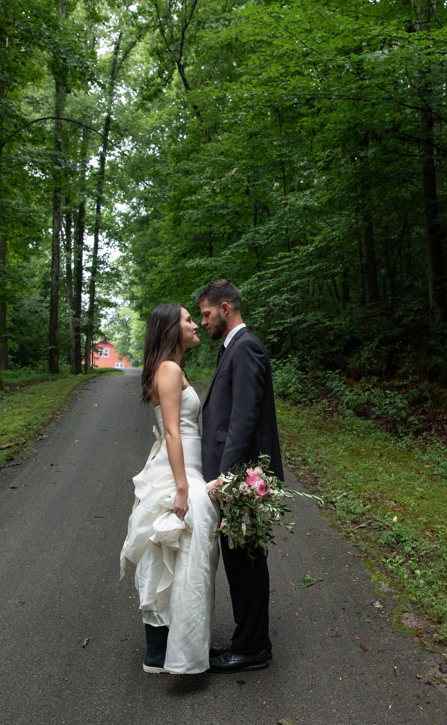 Photo of a bride and groom standing on a road lined by trees, looking at each other while the groom holds the bouquet. Elopement photography