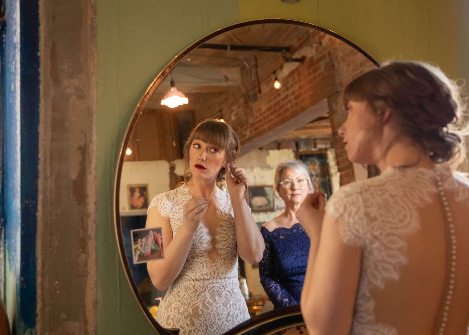 Photo of a bride looking in a mirror and putting on earrings, while the mother of the bride looks on in the background