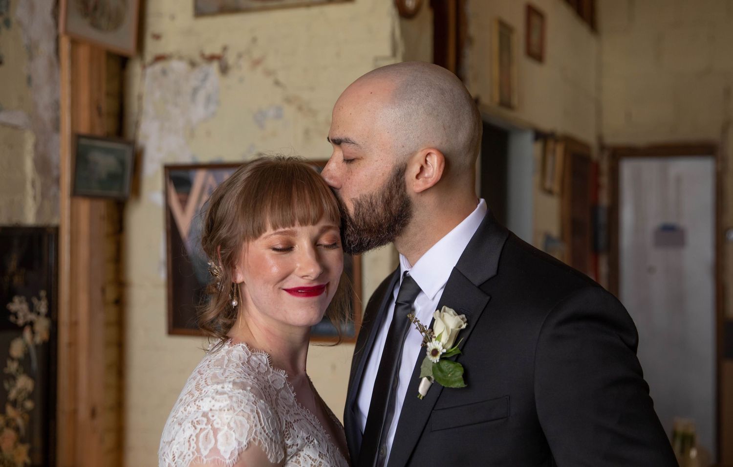 Photo of a bride and groom. She is looking at the camera, smiling with her eyes closed, and he is kissing the top of her head.
