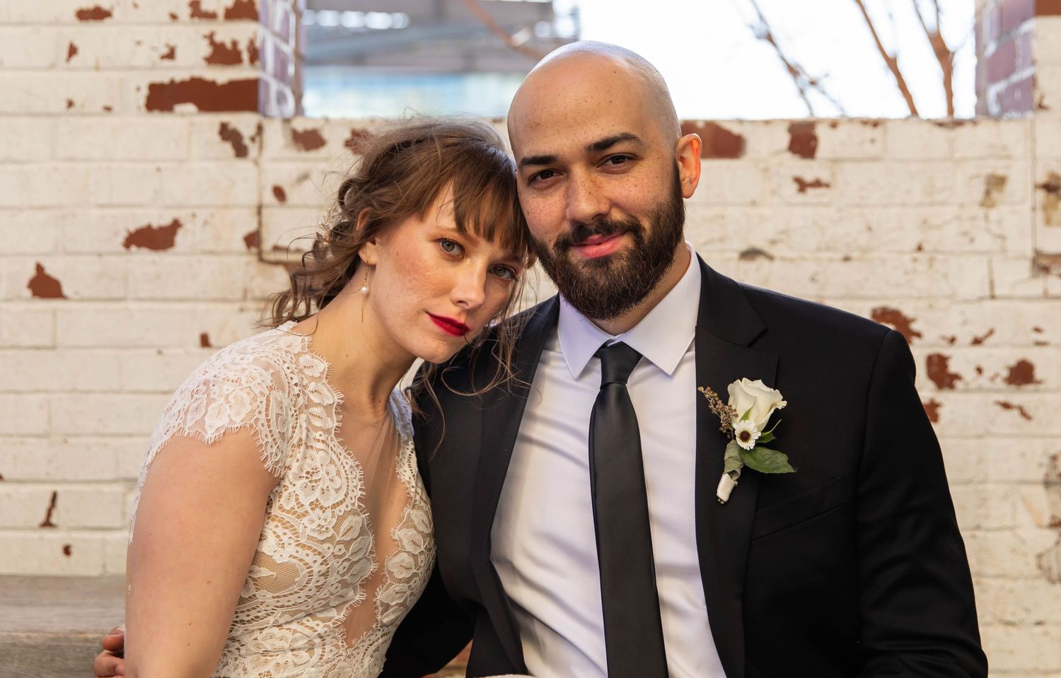 Photo of a bride and groom sitting in front of a white brick wall, leaning on each other and smiling. Raleigh, NC