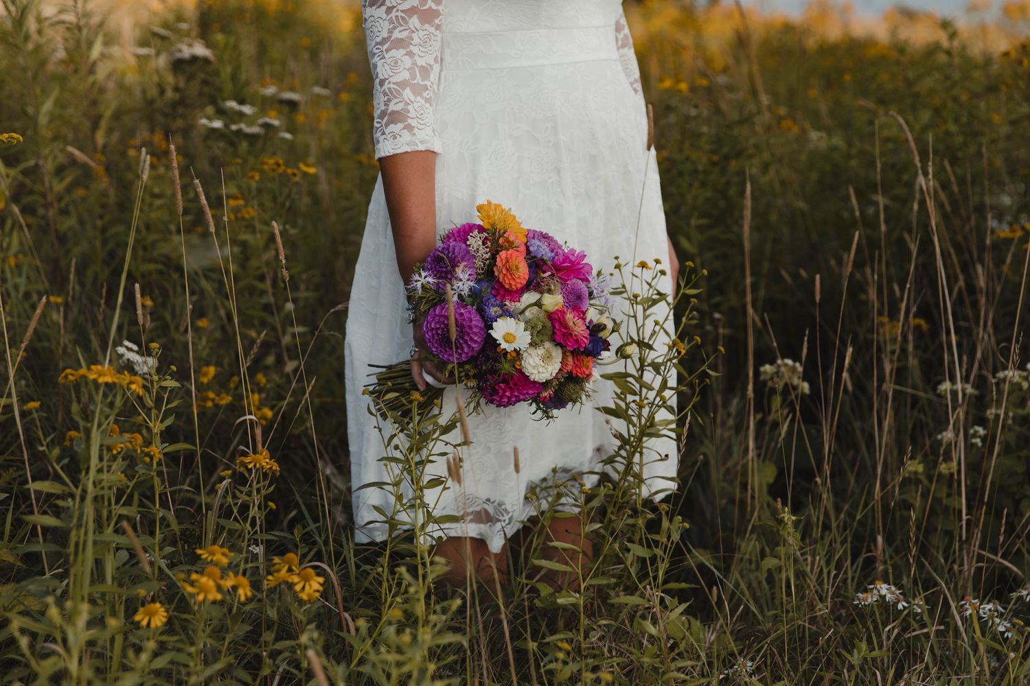 Photo of a bride standing in a field of tall grass and yellow flowers, holding her bouquet of flowers during an elopement Whitetop Mountain